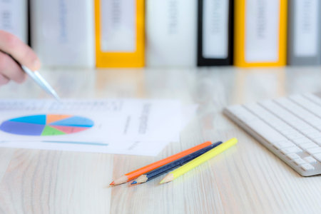 Manager working on presentation Side view of desk with keyboard, pencils, papers with coloured charts and human hand keeping pen. Stack of multicoloured office folders on the backgroundの写真素材
