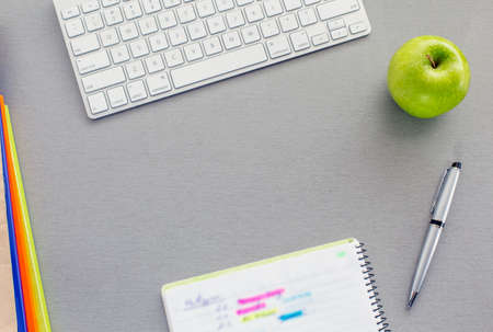 Office work space on grey desk with green apple From above view on grey wooden desk with well organised office supplies and green apple. Notepad with hand notes marked with bright coloursの写真素材