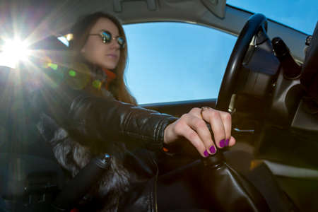 Confident female driver Young woman drives car, focus on her hand switching transmission grip. Steering wheel on the right, blues sky in the window, bright sunbeams on the leftの写真素材