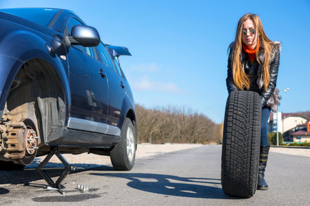 Female biker rolls big wheel Young female, dressed biker style, rolls big wheel towards broken car