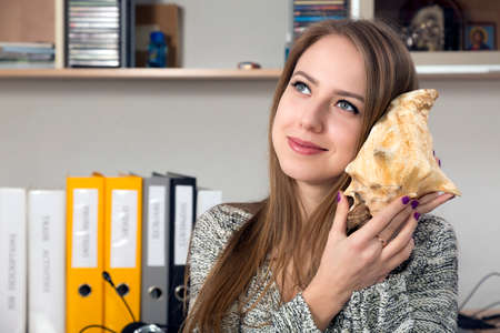 Beautiful smiling Caucasian young woman holding large sea shell and listening the sound dreamily looking into far Office interior casual clothingの写真素材