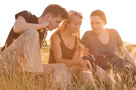 Family of three people sitting on grassy lawn watching on display of photo camera back light sun sunset sun on backgroundの写真素材