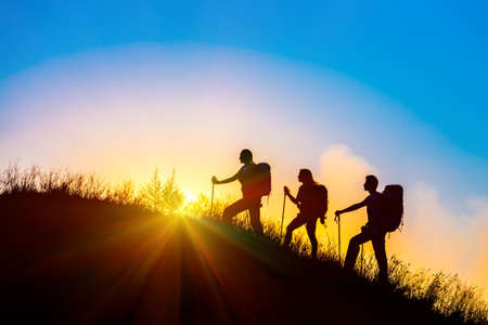 Group of people silhouettes walking toward mountain summit with backpacks hiking trekking gear meeting uprising sun sunbeams and blue sky of background