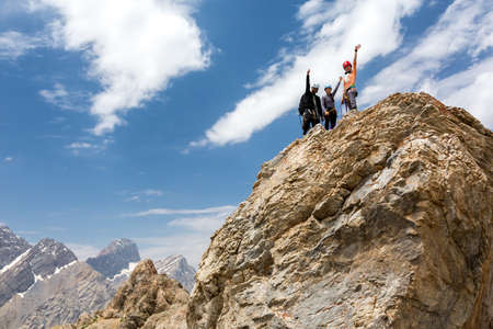 Happy people embracing raising hands up on pointed rocky cliff blue sky backgroundの写真素材