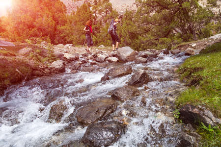 People going across mountain creek fast streaming water jumping on rocks green meadow and forest along river forest wild landscape and sun on backgroundの写真素材