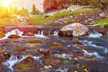 Rapid water stream of river on foreground and group of people hiker walking up hill footpath on background with bright sun shiningの写真素材