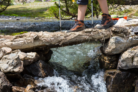 Close up of legs in heavy trekking boots walking with hiking sticks across rapid mountain river throw timber log primitive bridgeの写真素材