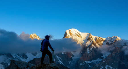 Silhouette of Athlete Walking on Rocky Peak holding Ice Axe and Rope Unusual Cloud Cover and Summits Illuminated by Rising Sun Backgroundの写真素材