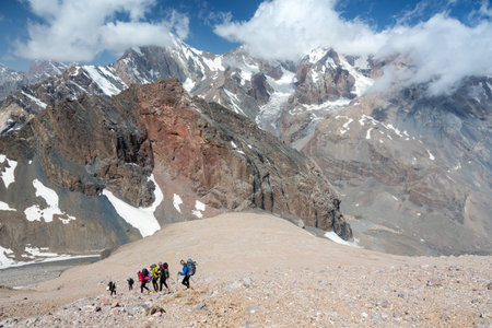 Group of People in Sporty Style Clothing with Backpacks and Walking Poles Mountain Landscape with Blue Sky and Clouds on Backgroundの写真素材