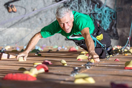 Portrait of Mature Male Climber Moving Up on Outdoor Climbing Wall Sporty Clothing on Fitness Training Intense but Positive Face Using Rope and Belaying Gearの写真素材