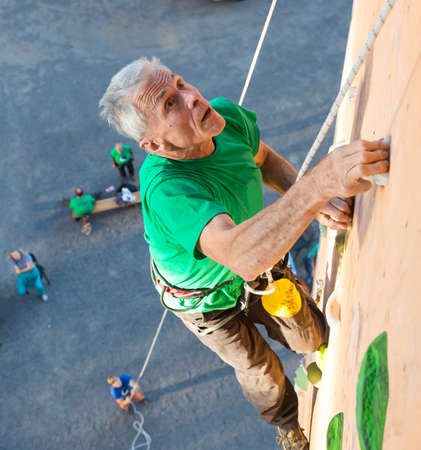 Elderly Male Climber Makes Hard Move and Looking High Up on Outdoor Climbing Wall Sport Competitions Very Emotional Face Belaying Partner Fans Staying Remote Groundの写真素材