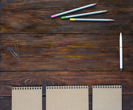 Top View of Natural Wood Background Line of Folded Notepads in Serene Beige White Pen and Three Colored Pencils Paper Metallic Clipsの写真素材