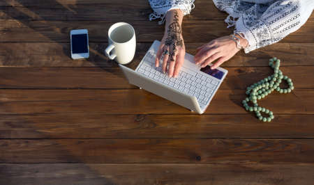 Natural Wooden Desk with White Computer Large Mug Telephone and Rosary Woman Typing on Keyboard with Stylish Tattoo on Wrist Top Viewの写真素材