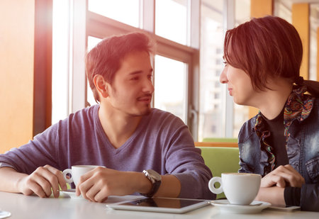 Young Couple Talking at Coffee Shop Sitting at White Desk with Tablet PC and Telephones on Table and...