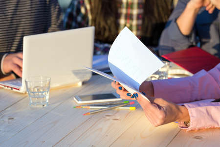 Hands of Businesswoman Holding Paper Prints of Presentation and Discussing with Partners Sitting around Wood Table with Laptop Pencils Telephone Glass of Water and Notepadsの写真素材
