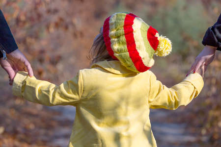 Two Generation Family Walking in Autumnal Forest Father Mother Holding Hands of Little Baby Girl Bright Yellow Clothing Coat and Cap Close Up Rear Viewの写真素材