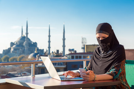 Traditionally dressed Muslim Woman working on computer at outdoor balcony with oriental urban landscape with minarets and blue sky on backgroundの写真素材