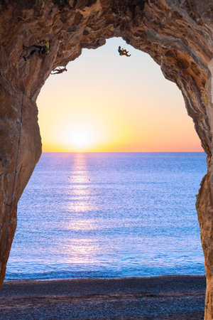 Deep high Rock Cave on Seafront and extreme Climbers Sea and Rising Sun on Backgroundの写真素材