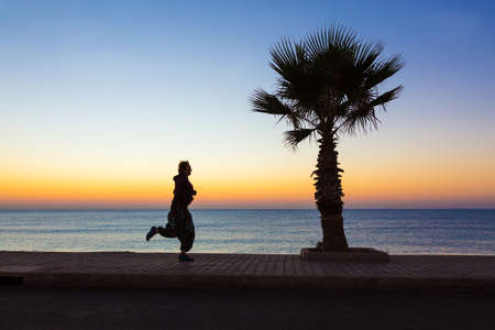Young Woman jogging on Seafront making Morning Fitness Tropical Palm Tree colorful Dawn blue Sea. Focus on Palm Tree and paved path, jogger body slightly blurred in moveの写真素材