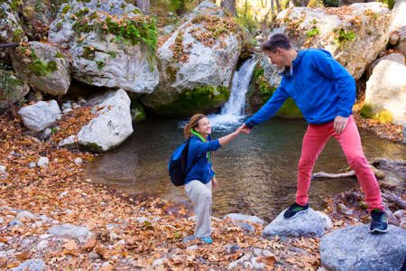 Two Hikers young Man and Smiling Woman holding hands helping each other walking mountain stream with waterfallの写真素材