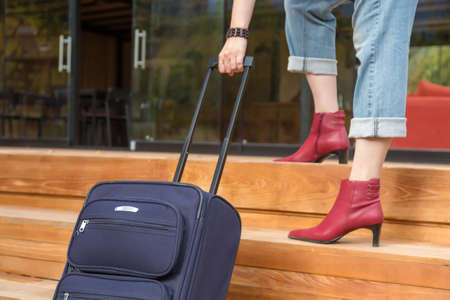 Person walking up on wooden Stairs pulling Travel Suitcase female Legs and Hand casual vacation Clothing red attractive high Heels Shoes and Jeansの写真素材