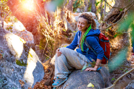 Cute smiling Girl with Day Backpack and sporty casual Clothing sitting on large fallen Tree Stalk in wild Forest Outdoors Sunlightの写真素材