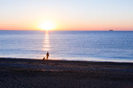 People traveling Beach Holiday Concept. Female Silhouette walking along Ocean Coast pulling Luggage Bag Sea and Sunrise on backgroundの写真素材