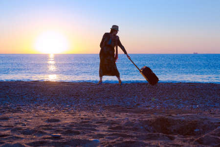 Silhouette of Lady walking along Sea Surf pulling Travel Suitcase Sunrise and Beach Vacation Conceptの写真素材