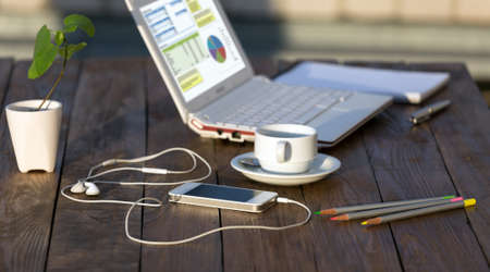 Side view of working Place on natural wooden Table with white Computer Telephone Coffee Mug Pencils Pen Notepad and Chart on Laptop Screenの写真素材