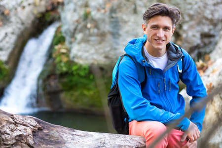 Portrait of handsome Male Hiker sitting on fallen Tree Trench at Mountain Lake sportive Clothing Jacket and Backpackの写真素材
