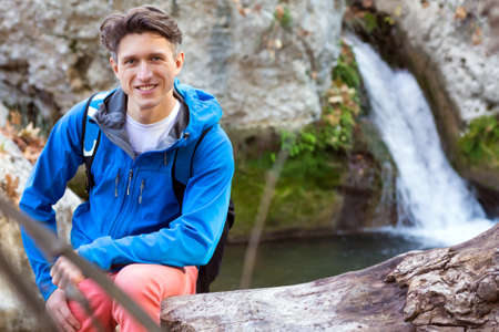 Portrait of handsome Male Hiker sitting on fallen Tree Trench at Mountain Lake sportive Clothing Jacket and Backpackの写真素材
