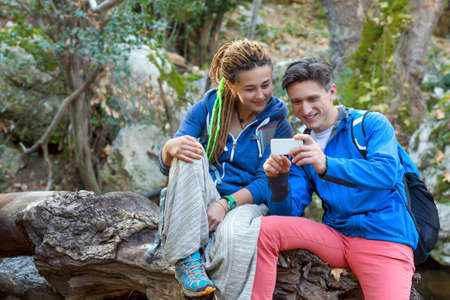 Man and Girl sitting on fallen Tree relaxing at Spring Forest and looking at Telephone Screen reviewing image taken smiling Faces Sportive Clothing Backpacksの写真素材