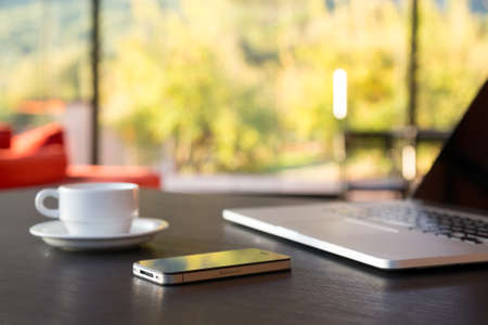 Business Travel Composition with cropped Computer Coffee Mug and Telephone on black grey Wood Table and large Windows Garden and Mountains on Backgroundの写真素材