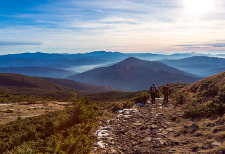 Two Hikers Man and Woman sportive Family walking along Mountains grassy Trail Scenic View of stacked smoky Ridges and Evening Sunlight on Backgroundの写真素材