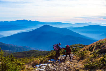 Sportive Couple on Hike in Mountains Man helping Woman holding her Hand stacked smoky Ridges and Sunlight on Backgroundの写真素材