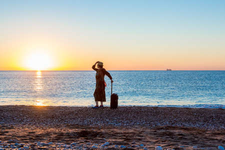 People traveling Beach Holiday Concept. Female Silhouette staying along Ocean Coast with Luggage Bag enjoying scenic View Sea and Sunrise on backgroundの写真素材
