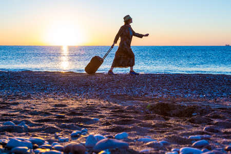 People traveling Beach Holiday Concept. Female Silhouette walking along Ocean Coast pulling Luggage Bag Sea and Sunrise on backgroundの写真素材