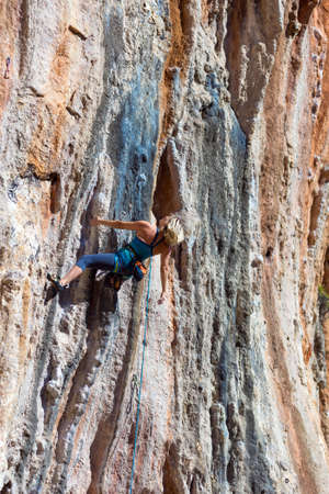 Blond Female Climber ascending Vertical Orange Rocky Wall unusual shape of Mountain Range on background Outdoor Sunshineの写真素材