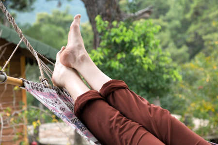 Person relaxing lying in Hammock at rural cottage garden female legs green flora and wooden hut on backgroundの写真素材