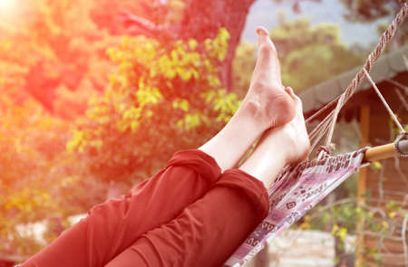 Person relaxing lying in Hammock at rural cottage garden female legs green flora and wooden hut on backgroundの写真素材