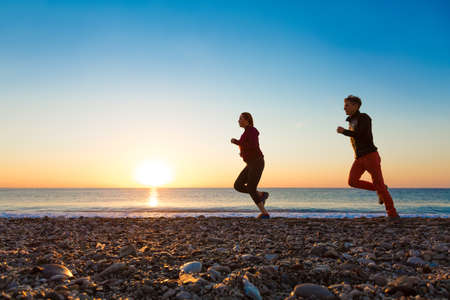 Sporty Couple Man and Woman doing Morning Jogging on Sea Beach at Bright Sunrise Silhouettesの写真素材