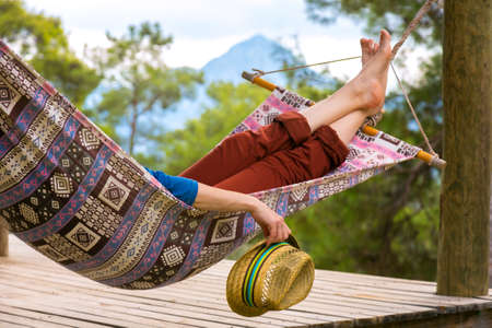 Person relaxing in Hummock holding Travel Hat in Hand at Summer Garden with Forest and Mountains on Background Landscapeの写真素材