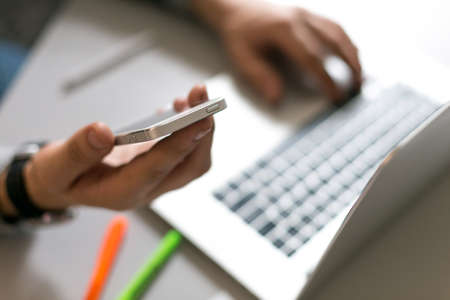 Man holding telephone and working on laptop at grey office table with pen and green and orange markersの写真素材
