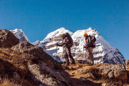 Two adult Mountain Climbers with Backpacks Gear and high Altitude Clothing walking on rocky Ridge with Himalaya Summits on Backgroundの写真素材