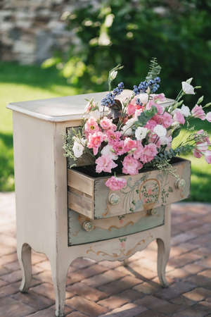 A vintage beige bedside table stands outside in the summer, decorated with a bouquet of pink roses flowersの写真素材