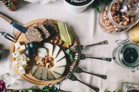 Sliced goat cheese with Dorblu with slices of bread, pears, grapes on a wooden plate. Nearby lie the fork, butter knife, a glass jar with hazelnuts. Top viewの写真素材