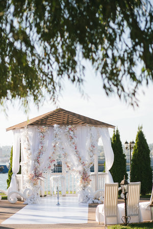 White wedding arch arbor decorated with white flowers and cloth, chairs, on the river bankの写真素材