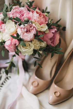A wedding bouquet of pink peonies, white roses, and eucalyptus, besides beige women's shoes, on which lie the wedding rings. background white clothの写真素材