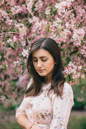 Portrait of a beautiful woman standing near a pink flowering apple tree. Looks down