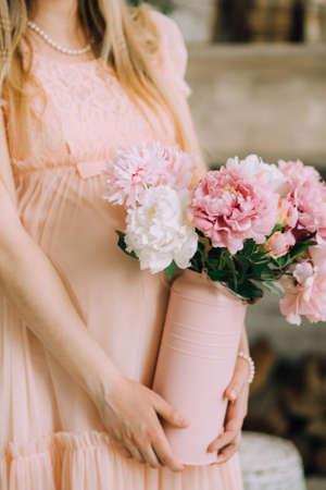 A pregnant woman in a delicate pink dress and a pearl necklace holds a vase of peonies with her handsの写真素材